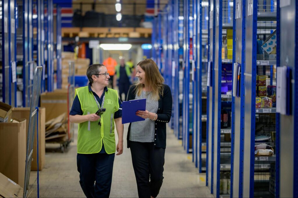 Two women warehouse employees walking down an aisle between shelves at a warehouse facility