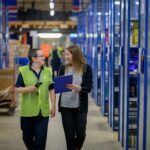 Two women warehouse employees speaking with one another as they walk down an aisle